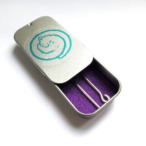 May include: A silver tin with a turquoise cat design on the lid. The tin is open and reveals a purple interior with two silver knitting needles.