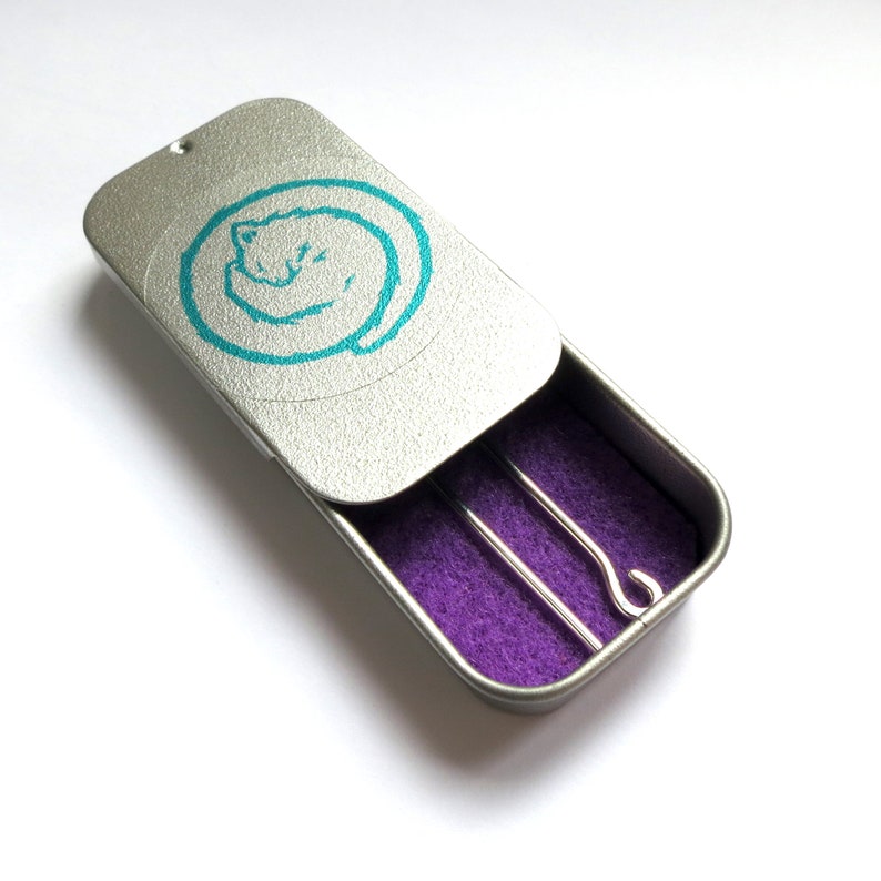 May include: A silver tin with a turquoise cat design on the lid. The tin is open and reveals a purple interior with two silver knitting needles.