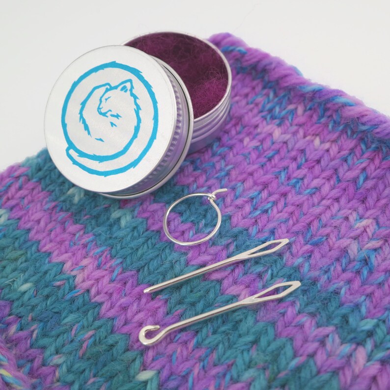 May include: A small silver tin with a blue cat design on the lid, a silver hoop earring, and two silver crochet hooks on a purple and blue striped knitted fabric.