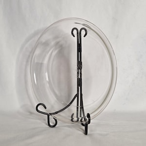 May include: A clear glass plate displayed on a black metal stand. The plate is round with a slightly raised edge. The stand has a curved design with a central vertical support and curved feet.