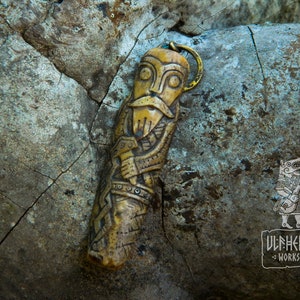 May include: A carved bone pendant depicting a Viking warrior with a beard and holding a weapon. The pendant is attached to a gold-colored metal loop. The pendant is resting on a textured, gray rock. The text "ULFHEDNAR WORKSHOP" is visible in the lower right corner of the image.