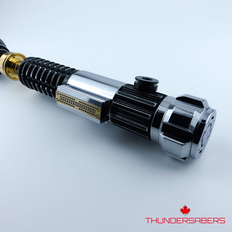 May include: A close-up of a lightsaber with a silver, black, and gold finish. The hilt has detailed designs and a cylindrical body. The lightsaber has a complex design with a detailed emitter and a textured grip.