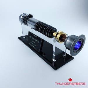 May include: A detailed lightsaber prop in silver and black, presented on a black stand. The lightsaber features a gold accent and a blue glowing emitter. The stand includes a plaque that reads "Obi-Wan Kenobi".