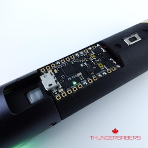 Puede incluir: Primer plano de un dispositivo electr&oacute;nico negro con componentes de placa de circuito expuestos. La placa tiene varios chips, puertos y el texto "PB V3.91". El dispositivo tiene un exterior negro texturizado y el logotipo "THUNDERSABERS" con una hoja de arce.