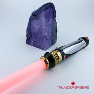 Puede incluir: Un sable de luz negro y plateado con una hoja roja, detalles dorados y un bot&oacute;n rojo. Una geoda morada grande en el fondo. El texto "THUNDERSABERS" est&aacute; en la parte inferior.
