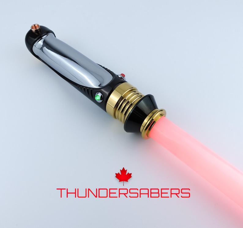 Puede incluir: Un sable de luz con un mango cromado y negro, detalles dorados y una hoja rosa brillante. El mango tiene un bot&oacute;n rojo y verde. El texto "THUNDERSABERS" se muestra debajo con una hoja de arce roja.