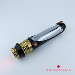 Puede incluir: Un sable de luz negro y dorado con una hoja cromada. El mango presenta una secci&oacute;n dorada, un bot&oacute;n rojo y un bot&oacute;n verde. El texto "THUNDERSABERS" y un logotipo de hoja de arce son visibles.