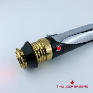 Puede incluir: Un sable de luz con un dise&ntilde;o negro, dorado y plateado. El mango presenta detalles dorados y un bot&oacute;n rojo. La hoja no es visible. El texto "THUNDERSABERS" est&aacute; en la parte inferior.
