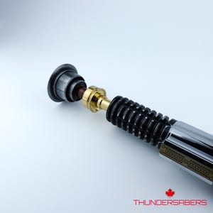 May include: A lightsaber in black, silver, and gold. The hilt features a ribbed black grip, a gold midsection, and a silver blade emitter. The text "THUNDERSABERS" is visible at the bottom of the image.