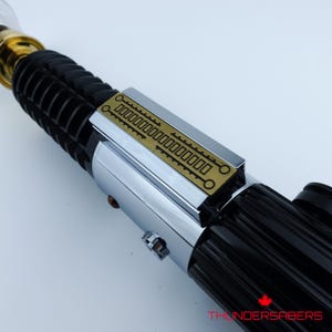 May include: A close-up of a lightsaber with a black handle and a silver and gold hilt. The lightsaber has a gold-colored section near the top and a rectangular gold plate with etched details. The text "THUNDERSABERS" is visible.