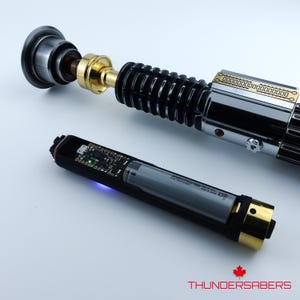 May include: A lightsaber with a black, silver, and gold design is shown. The hilt has detailed features, and a separate component with a circuit board and battery is visible. The logo "THUNDERSABERS" is at the bottom of the image.