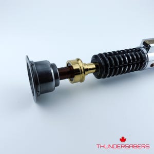 May include: A detailed lightsaber prop with a silver, black, and gold design. The hilt features ribbed black sections, a gold midsection, and a silver emitter. The text "THUNDERSABERS" is at the bottom.