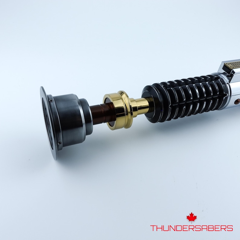May include: A detailed lightsaber prop with a silver, black, and gold design. The hilt features ribbed black sections, a gold midsection, and a silver emitter. The text "THUNDERSABERS" is at the bottom.
