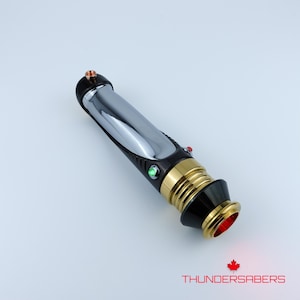 Puede incluir: Un sable de luz plateado y negro con detalles dorados y botones de activaci&oacute;n rojos y verdes. La base del mango tiene un brillo rojo. El texto "THUNDERSABERS" est&aacute; impreso en la parte inferior.