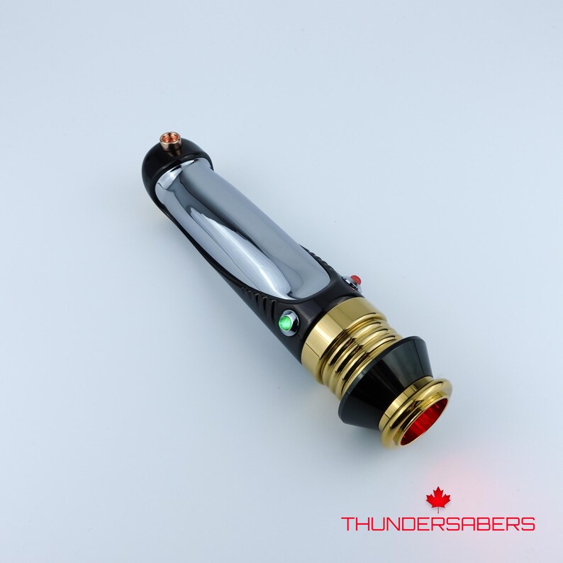 Puede incluir: Un sable de luz plateado y negro con detalles dorados y botones de activaci&oacute;n rojos y verdes. La base del mango tiene un brillo rojo. El texto "THUNDERSABERS" est&aacute; impreso en la parte inferior.