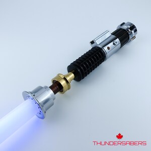 May include: A silver and black lightsaber with a glowing white and blue blade. The hilt features a black ribbed grip, silver accents, and a gold connector. The text "THUNDERSABERS" is in red.