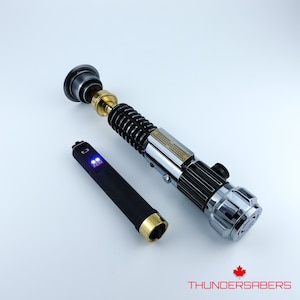 May include: A detailed lightsaber prop with a black, silver, and gold finish. The hilt features intricate detailing, and a separate black control unit with blue lights is included. The Thundersabers logo is visible in the bottom right corner.
