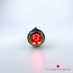 Puede incluir: Una empu&ntilde;adura de sable de luz negra y dorada con un emisor rojo iluminado. El emisor presenta un dise&ntilde;o circular con m&uacute;ltiples luces LED rojas brillantes. El texto "THUNDERSABERS" se muestra en blanco, con un logotipo de hoja de arce roja.