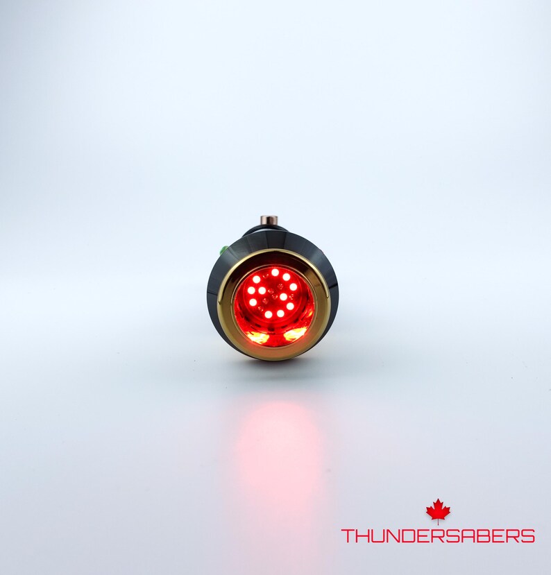 Puede incluir: Una empu&ntilde;adura de sable de luz negra y dorada con un emisor rojo iluminado. El emisor presenta un dise&ntilde;o circular con m&uacute;ltiples luces LED rojas brillantes. El texto "THUNDERSABERS" se muestra en blanco, con un logotipo de hoja de arce roja.