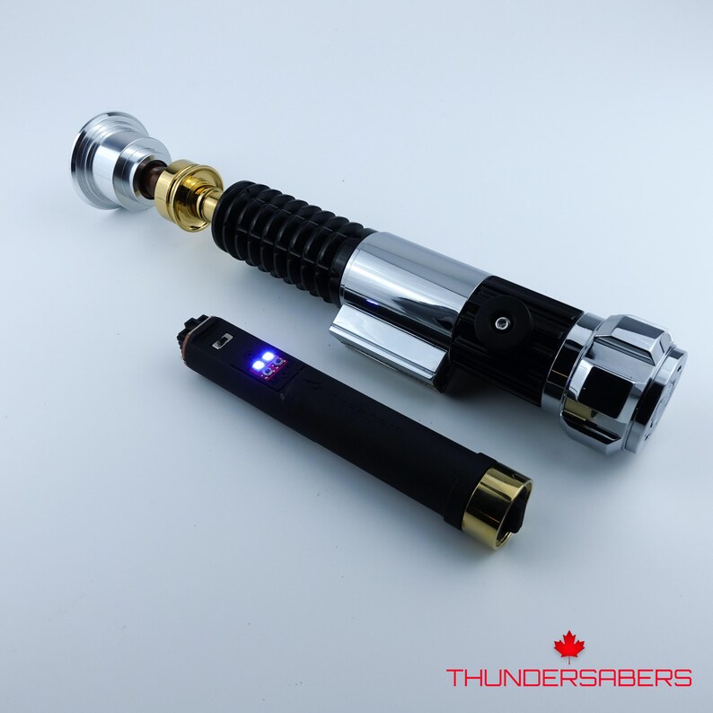 May include: A silver, black, and gold lightsaber prop with a black handle and a separate black device with blue lights. The lightsaber has a detailed design with a metallic finish. The prop is designed to resemble a lightsaber.