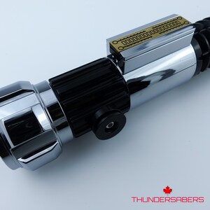 May include: A chrome and black lightsaber with a gold accent plate. The lightsaber has a detailed design with a cylindrical body and a textured grip. The Thundersabers logo is visible in the bottom right corner.