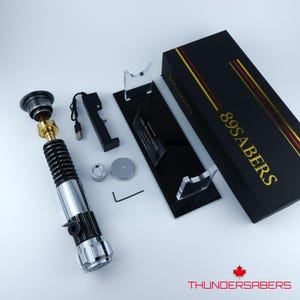 May include: A lightsaber prop with a silver, black, and gold finish, presented with a black display stand and a black box with gold lettering reading "89SABERS". Accessories include a charging cable and small metal components.