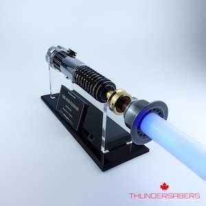 May include: A replica lightsaber, resembling Obi-Wan Kenobi's, with a silver and black handle and a glowing blue blade. The lightsaber is displayed on a black stand, with the text "OBI-WAN KENOBI" on a plaque. The overall design is highly detailed.