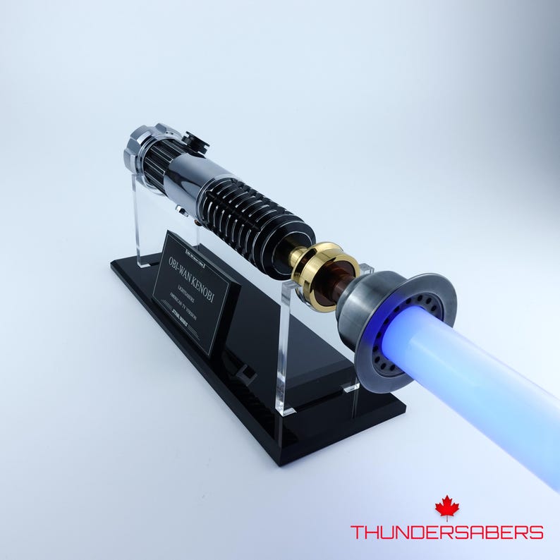 May include: A replica lightsaber, resembling Obi-Wan Kenobi's, with a silver and black handle and a glowing blue blade. The lightsaber is displayed on a black stand, with the text "OBI-WAN KENOBI" on a plaque. The overall design is highly detailed.