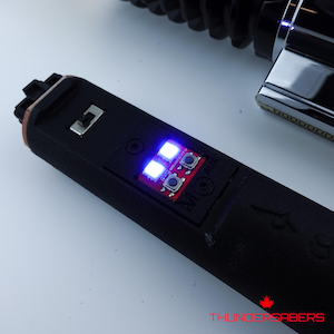May include: Close-up of a black lightsaber hilt with a red circuit board featuring two blue LED lights and two buttons. The text "THUNDERSABERS" is visible at the bottom.