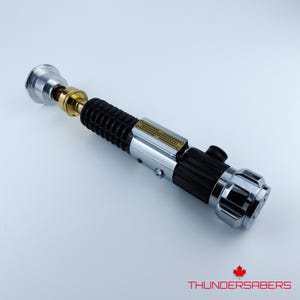 May include: A detailed lightsaber prop with a silver, black, and gold design. The hilt features a ribbed black grip, a silver midsection, and a gold and silver emitter. The end cap is chrome. The text "THUNDERSABERS" is at the bottom.