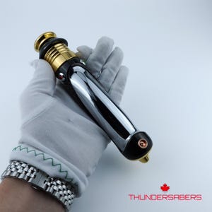 Puede incluir: Un sable de luz cromado y negro con detalles dorados. El mango presenta una superficie pulida y reflectante y un emisor dorado. El sable de luz se sostiene en una mano enguantada de blanco. El texto "THUNDERSABERS" es visible.