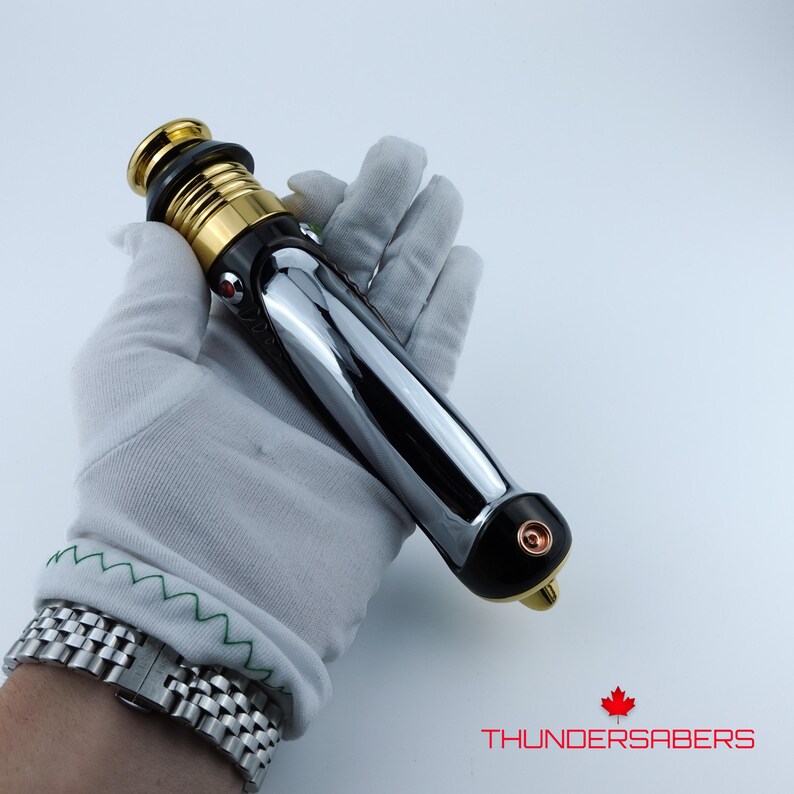 Puede incluir: Un sable de luz cromado y negro con detalles dorados. El mango presenta una superficie pulida y reflectante y un emisor dorado. El sable de luz se sostiene en una mano enguantada de blanco. El texto "THUNDERSABERS" es visible.