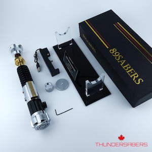 May include: A disassembled lightsaber with a black and silver handle, gold accents, and a silver top. Accessories include a black stand, a black box with gold lettering, and a small silver and black charging component. The box reads "89SABERS".