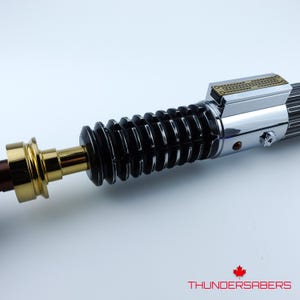 May include: A close-up of a lightsaber with a black ribbed handle, gold detailing, and a silver metallic top. The lightsaber features detailed craftsmanship and a shiny finish. The brand name "THUNDERSABERS" is displayed at the bottom.