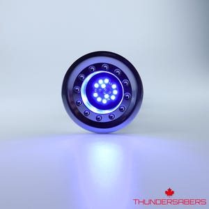 May include: A circular, metallic blue device with a central cluster of bright blue LED lights. The device has a ring of smaller lights around the central cluster. The text "THUNDERSABERS" and a maple leaf are visible at the bottom.