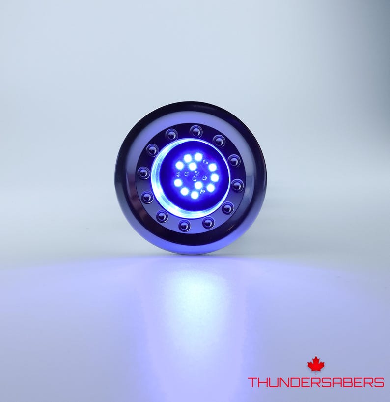 May include: A circular, metallic blue device with a central cluster of bright blue LED lights. The device has a ring of smaller lights around the central cluster. The text "THUNDERSABERS" and a maple leaf are visible at the bottom.
