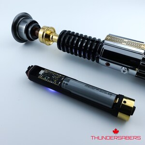 May include: A lightsaber with a black, silver, and gold design is shown. The hilt has a ribbed section and detailed features. A separate black component with a battery is also visible. The text "THUNDERSABERS" is at the bottom.