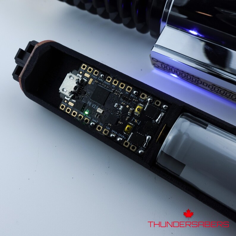 May include: A detailed view of a lightsaber hilt, revealing its internal components. The black housing is open, displaying a circuit board with various electronic components, a battery, and a USB port. The hilt's polished metal end glows with blue light. The text "THUNDERSABERS" is visible.