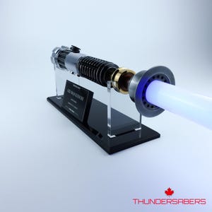 May include: Obi-Wan Kenobi's lightsaber replica, with a black and silver hilt and gold details. The lightsaber emits a bright blue blade. It is displayed on a black stand with a nameplate.