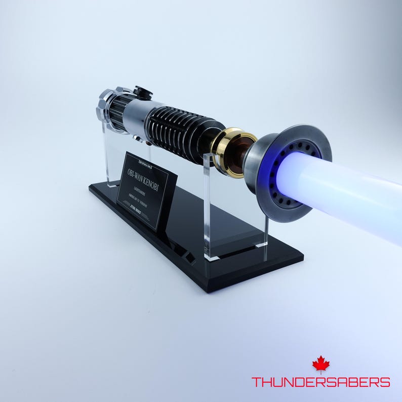 May include: Obi-Wan Kenobi's lightsaber replica, with a black and silver hilt and gold details. The lightsaber emits a bright blue blade. It is displayed on a black stand with a nameplate.