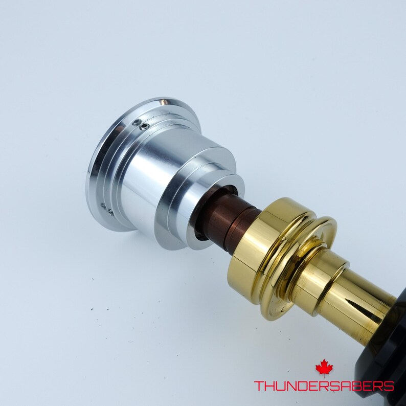 May include: A metallic lightsaber hilt with silver, gold, and brown components. The hilt has a detailed design with a Canadian maple leaf logo and the text "THUNDERSABERS". The lightsaber is set against a white background.