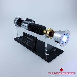 May include: Obi-Wan Kenobi lightsaber replica, featuring a black ribbed handle, silver and gold accents, and a glowing blue emitter. The lightsaber is displayed on a black stand with a nameplate.