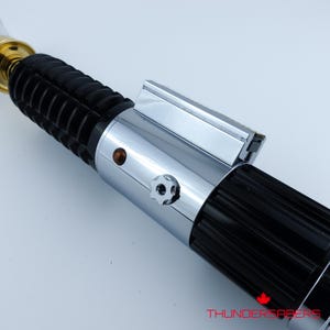 May include: A close-up of a lightsaber with a black ribbed handle and a silver metallic midsection. The lightsaber has a gold-colored emitter and a clear blade. The text "THUNDERSABERS" is visible in the bottom right corner.
