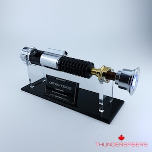 May include: A silver, black, and gold lightsaber prop, displayed on a black stand with a clear acrylic holder. The lightsaber has a ribbed black handle and a silver emitter. The stand has a plaque that reads "Obi-Wan Kenobi".