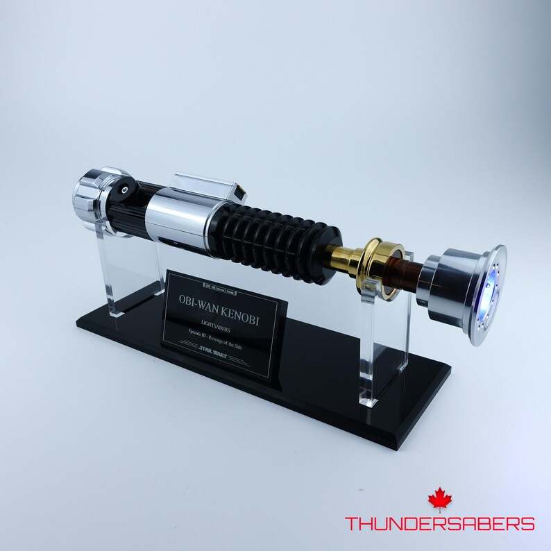 May include: A silver, black, and gold lightsaber prop, displayed on a black stand with a clear acrylic holder. The lightsaber has a ribbed black handle and a silver emitter. The stand has a plaque that reads "Obi-Wan Kenobi".