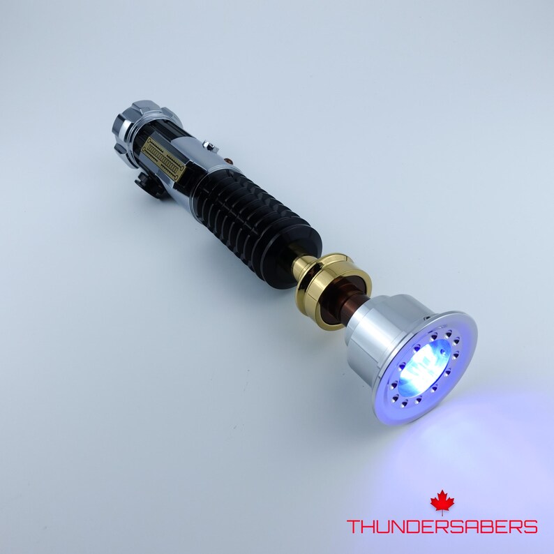 May include: A silver and black lightsaber with a black grip, gold accents, and a glowing blue emitter. The lightsaber has the text "THUNDERSABERS" and a maple leaf logo.