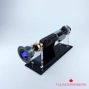 May include: A lightsaber with a blue blade, presented on a black stand. The hilt is a mix of silver, black, and gold. The text "THUNDERSABERS" is visible in the bottom right corner.