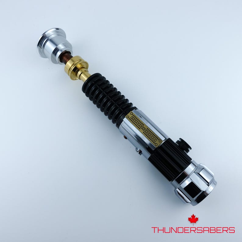 May include: A silver, black, and gold lightsaber prop with a ribbed black grip. The lightsaber has a silver emitter and pommel, with gold and brown accents. The text "THUNDERSABERS" is at the bottom.