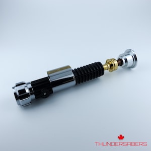 May include: A silver, black, and gold lightsaber prop with a ribbed black grip. The lightsaber has a cylindrical design with detailed metallic accents and a small rectangular plate. The text "THUNDERSABERS" is visible.