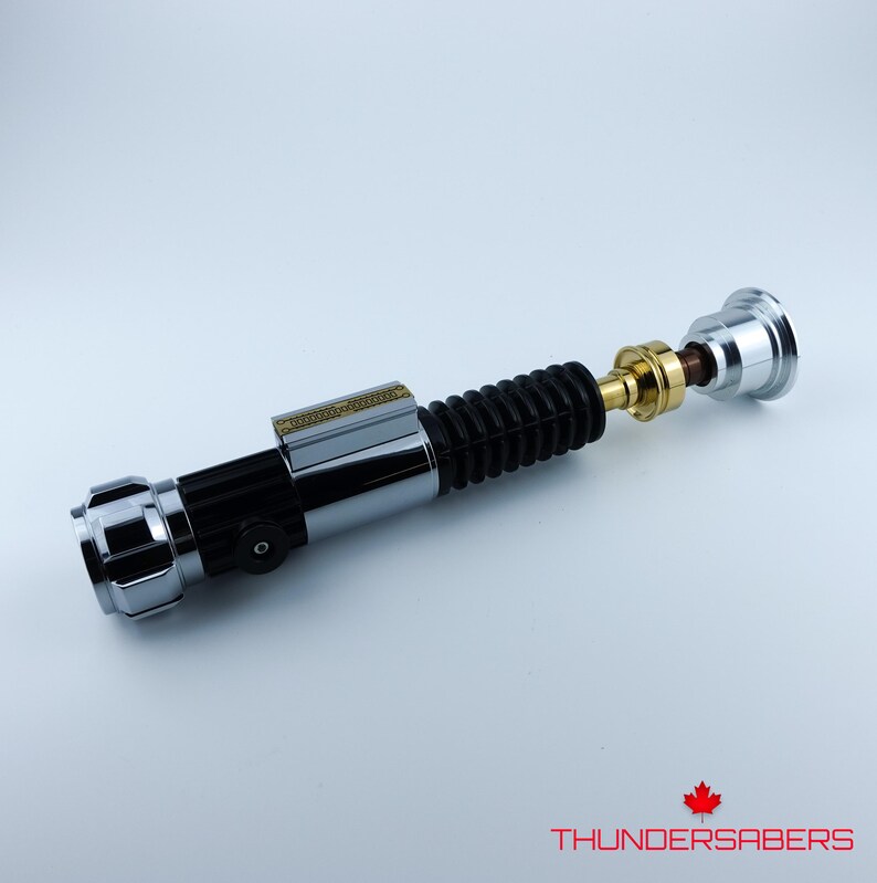 May include: A silver, black, and gold lightsaber prop with a ribbed black grip. The lightsaber has a cylindrical design with detailed metallic accents and a small rectangular plate. The text "THUNDERSABERS" is visible.